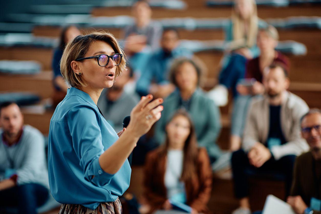 A woman with glasses stands before an audience, presenting educational material to her fellow students.